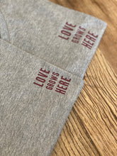 Load image into Gallery viewer, love-grows-here-slogan-on-grey-folded-tshirts
